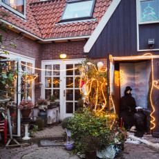 A costumed figure in a cloak beside a lit barn door in a De Rijp back garden