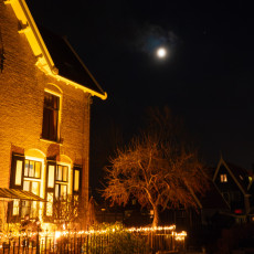 A full moon above a golden-lit house and candlelit railing in De Rijp