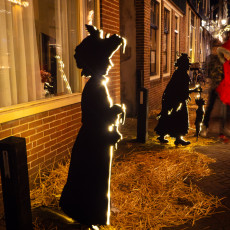 Lit silhouettes of Victorian figures standing on straw along a De Rijp street