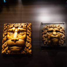 Two carved and gilded lion masks from ships, mounted on a dark gallery wall