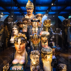 Rows of painted figureheads crowded together in a glass case, lit from above