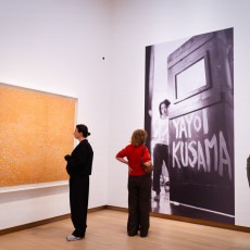 Visitors in front of a big orange net painting beside a photograph of Yayoi Kusama