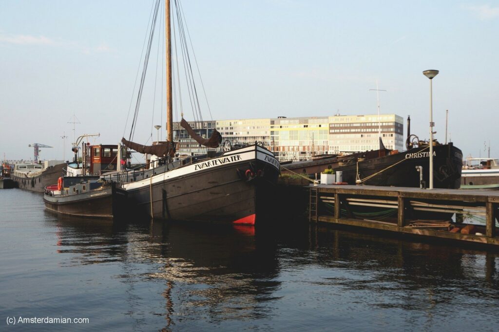 Amsterdam's Oude Houthaven features moored wooden ships named "Zwaarewater" and "Christina," with a modern building in the background under a clear sky