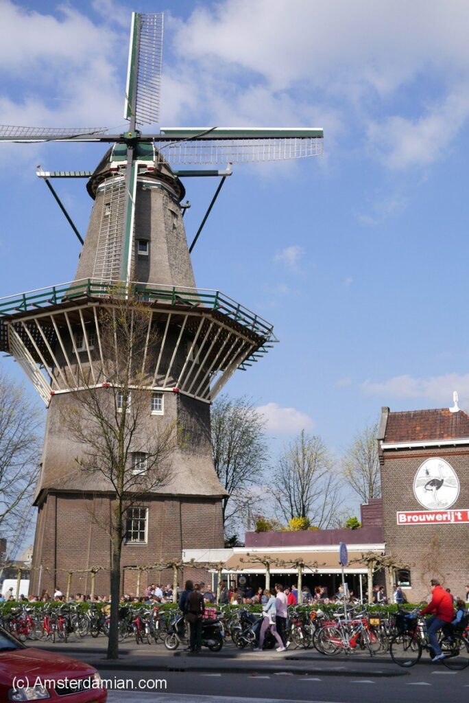 A bustling street scene in Amsterdam features a historic windmill, bicycles, and people near the IJ Brewery under a bright blue sky with scattered clouds
