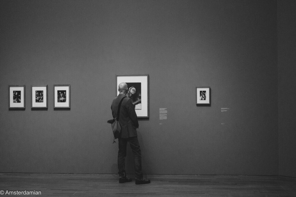 A man leans in close to look at a small framed black-and-white photograph on a grey wall at the Ed van der Elsken exhibition in the Rijksmuseum.