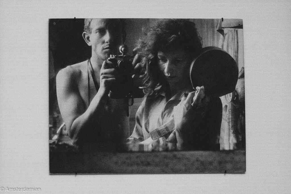 A large black-and-white photograph of a man holding a camera to his eye beside a woman with a round mirror, shown on a white gallery wall.