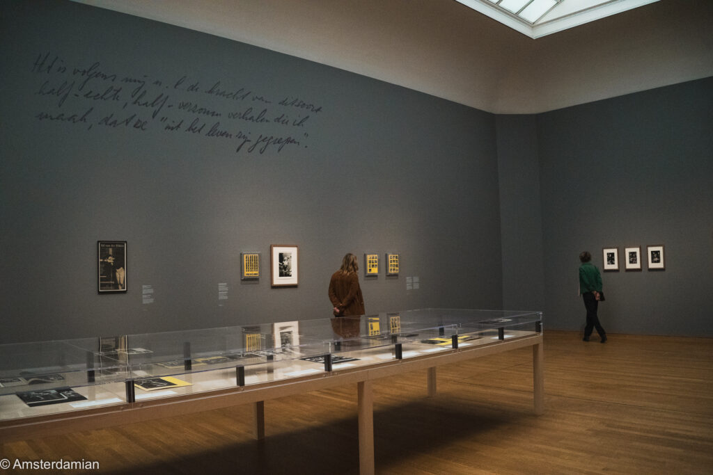 A Rijksmuseum gallery with a long glass display case, framed photographs and a large handwritten Dutch quotation on the wall, two visitors looking around.
