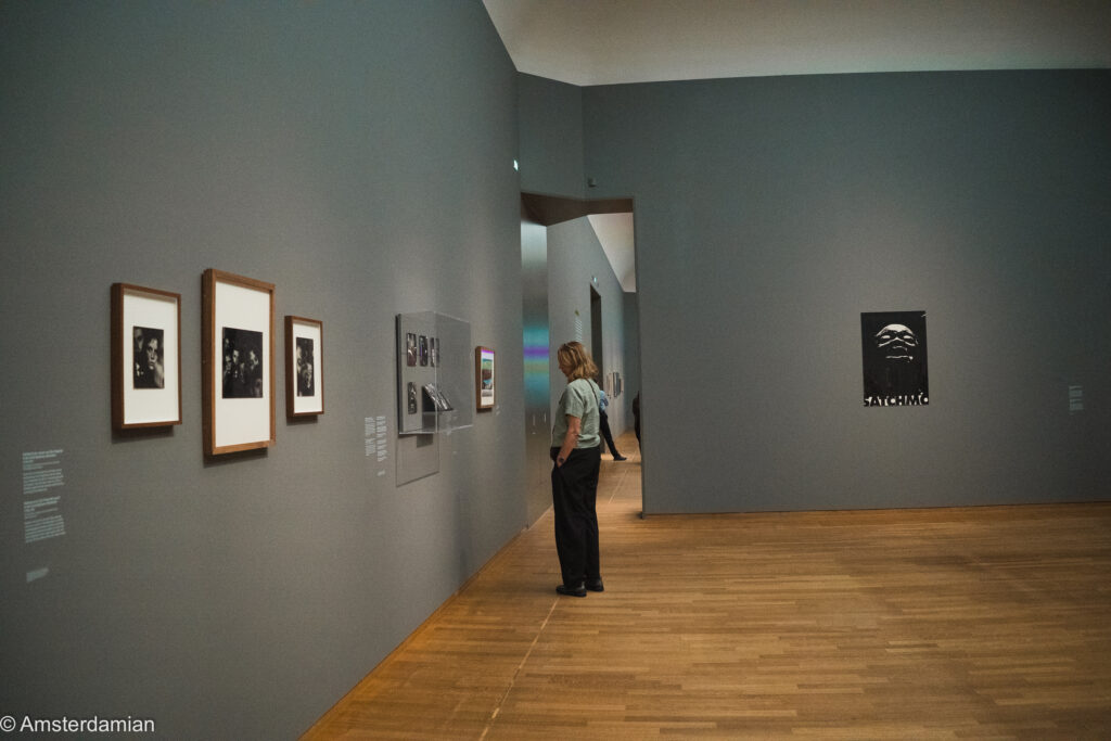 A visitor looks at framed photographs in a grey gallery, with Ed van der Elsken's black-and-white Satchmo poster on the far wall.