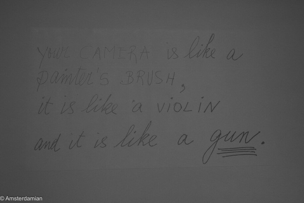 A handwritten quotation on a dark gallery wall reading 'your camera is like a painter's brush, it is like a violin and it is like a gun.'