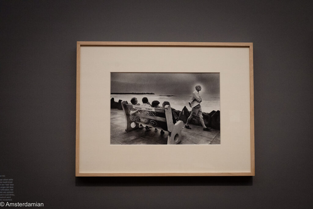 A framed black-and-white Van der Elsken photograph of people on a seafront bench as a boy walks past, hung on a dark gallery wall.