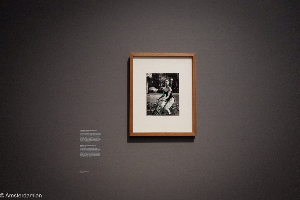 A framed Van der Elsken photograph of a woman cycling through Amsterdam in 1965, hung alone on a dark grey wall beside its label.