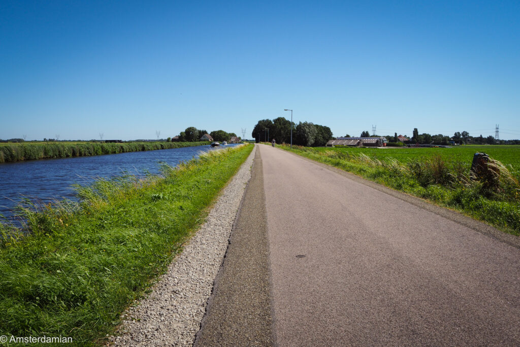 An empty asphalt cycle path running beside a canal through flat green polder under a clear blue sky north of Amsterdam
