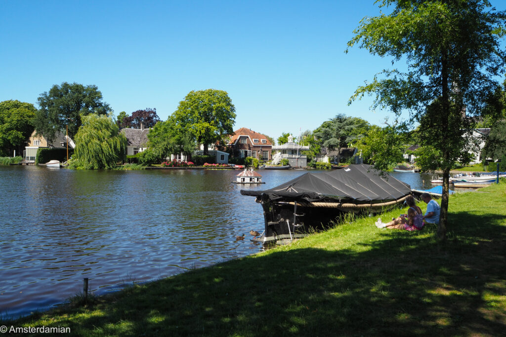 A woman and child sitting on the grass beside a lake and a tarp-covered wooden boat, with traditional Dutch houses and willow trees on the far bank