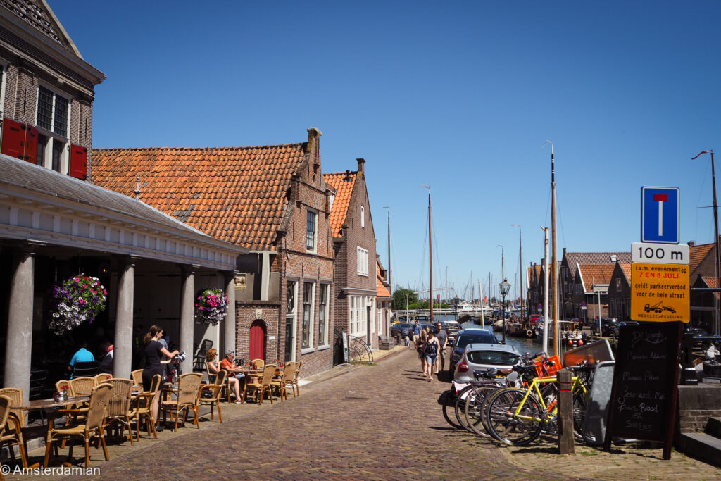 A cobbled harbour street with a cafe terrace under a colonnade, a stepped-gable brick house, parked bikes and traditional sailing boats moored along the quay