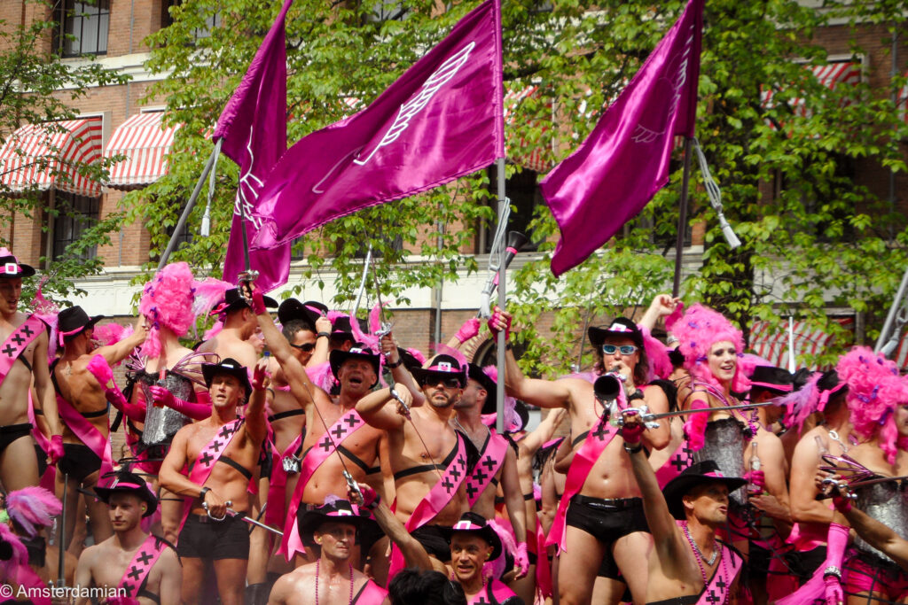 Men in pink and black cowboy outfits with Amsterdam sashes and magenta flags on a parade boat