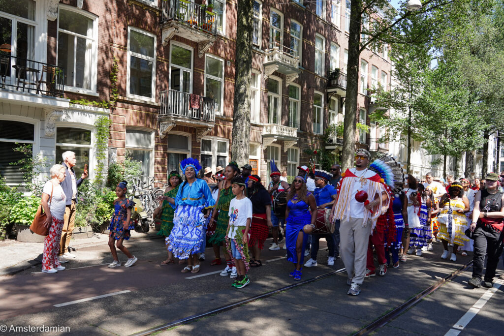 Bigi Spikri parade on an Amsterdam street lined with canal houses, participants in blue and green koto dress
