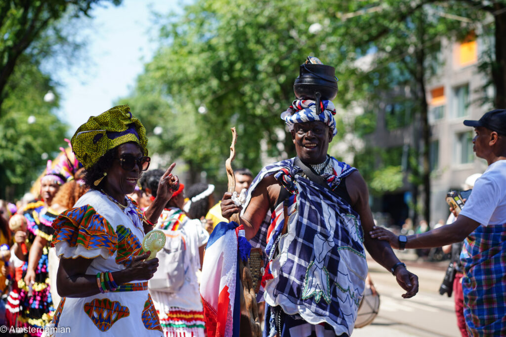 Two people in traditional Surinamese dress at Amsterdam's Bigi Spikri parade, one balancing a pot on his head
