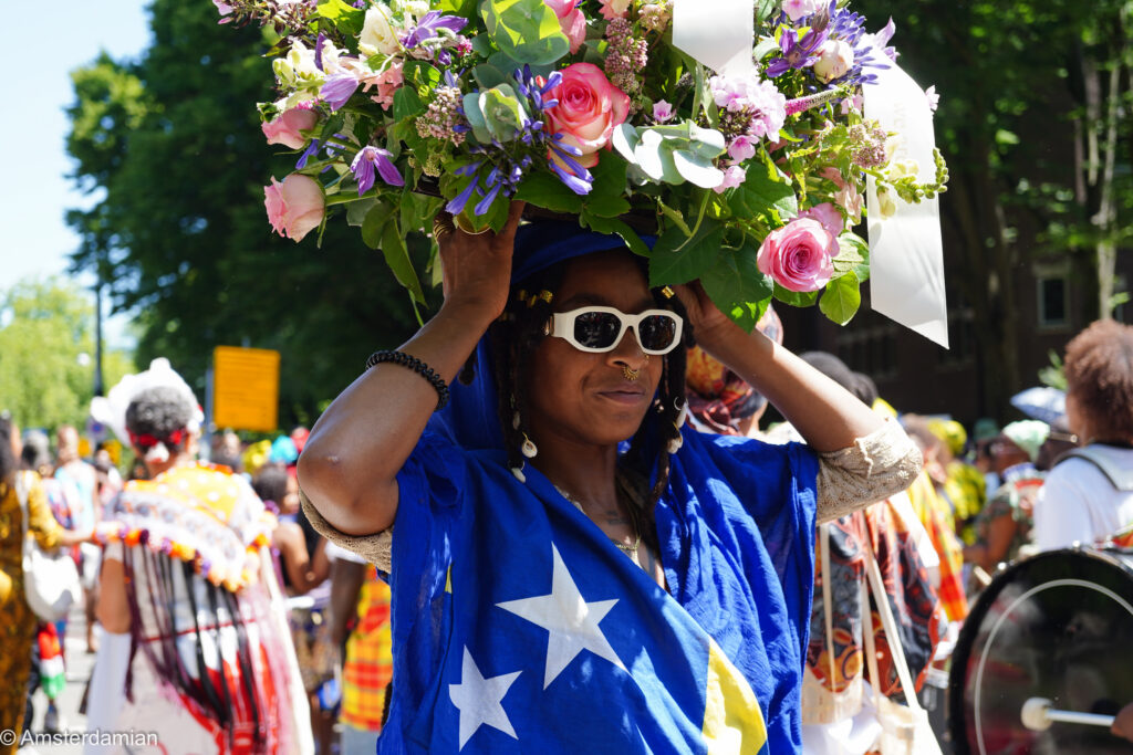 Parade participant balancing a basket of flowers on her head at Amsterdam's Bigi Spikri parade