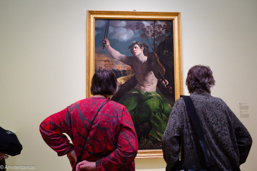 Two visitors in front of Dosso Dossi's painting of Apollo, crowned with laurel and raising a violin bow