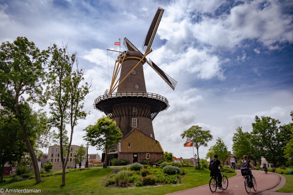 Molen de Valk, Leiden's tall windmill museum, with cyclists passing on the path below a cloudy sky