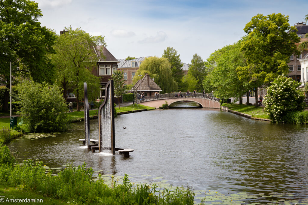 A leafy canal with a modern water sculpture and an arched footbridge on a sunny day in the Netherlands