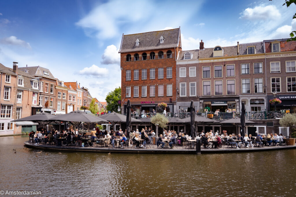Busy canalside terrace full of people dining under umbrellas beneath tall Dutch canal houses