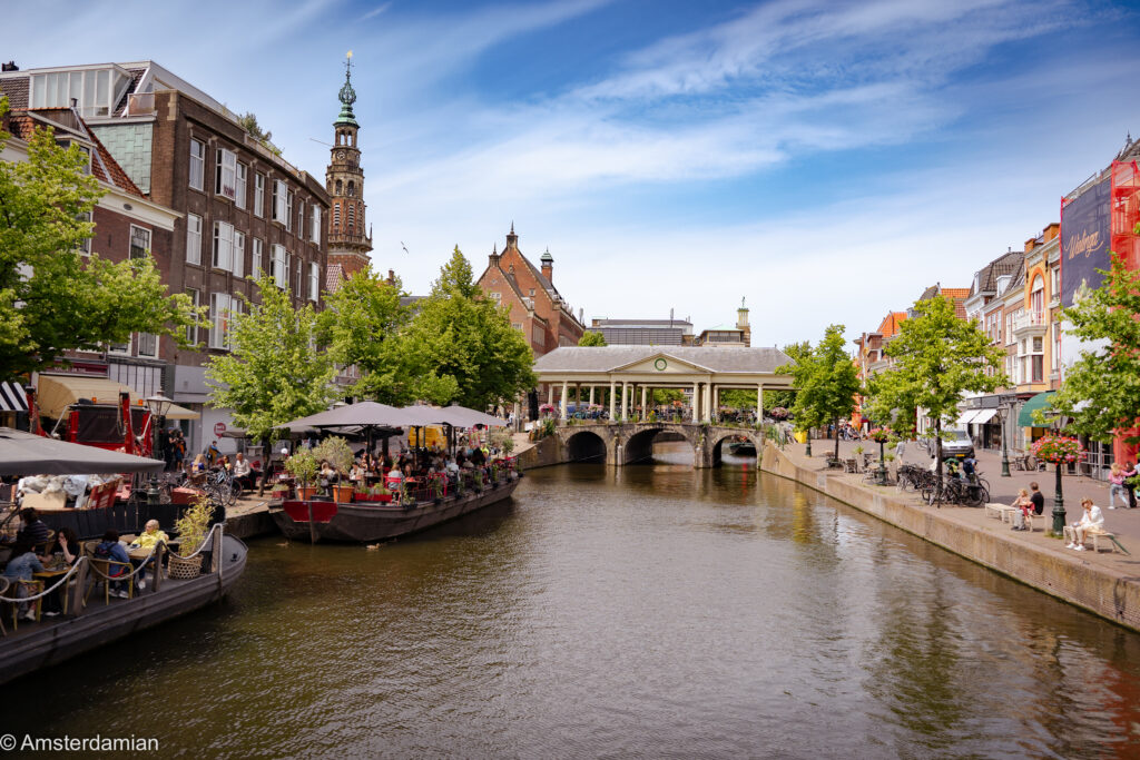 Sunny canal lined with cafe terraces and boats, a historic tower and covered bridge in the distance